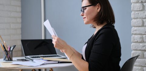 Woman wearing glasses reviewing paperwork while sitting at a desk with a laptop.