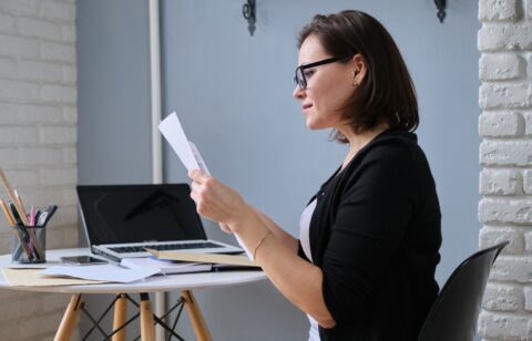 Woman wearing glasses reviewing paperwork while sitting at a desk with a laptop.