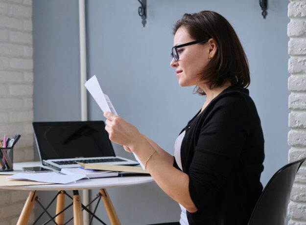 Woman wearing glasses reviewing paperwork while sitting at a desk with a laptop.
