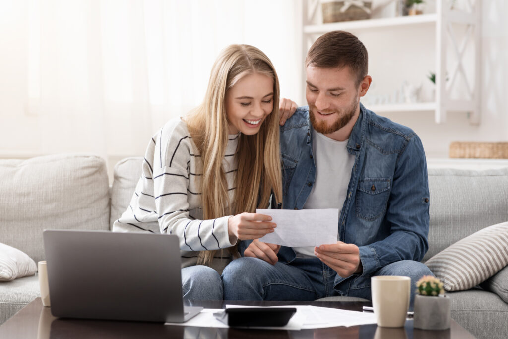 Couple smiling while reviewing paperwork together on a couch with a laptop.