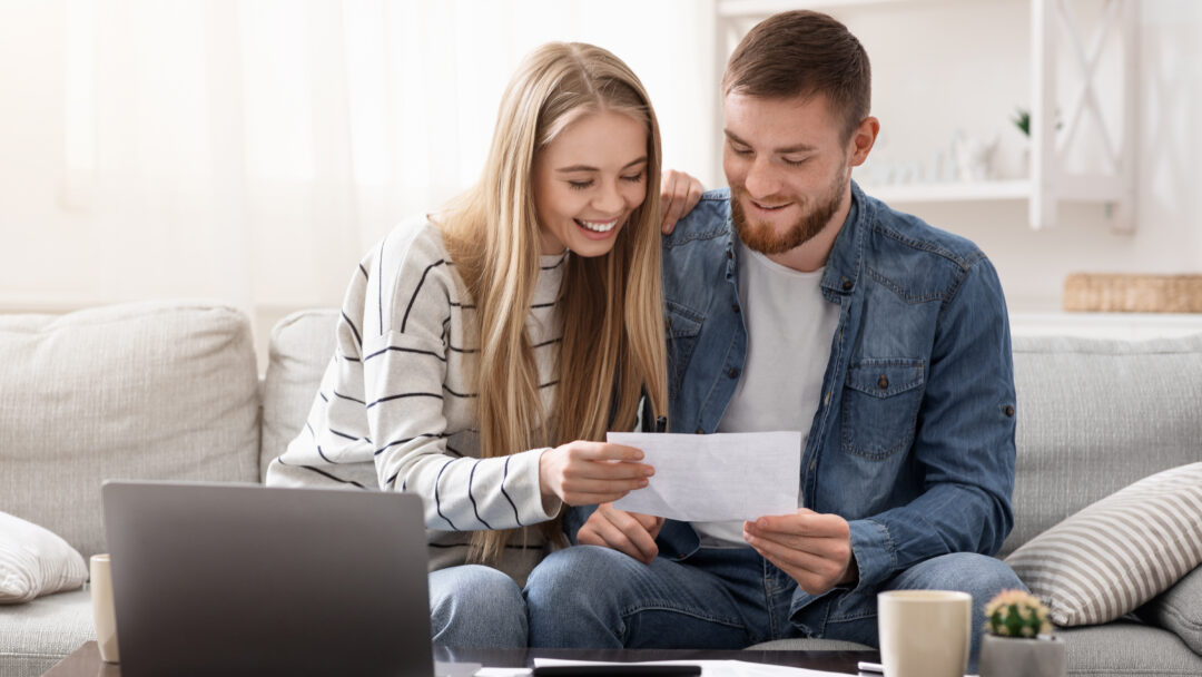 Couple smiling while reviewing paperwork together on a couch with a laptop.
