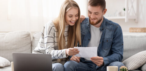 Couple smiling while reviewing paperwork together on a couch with a laptop.
