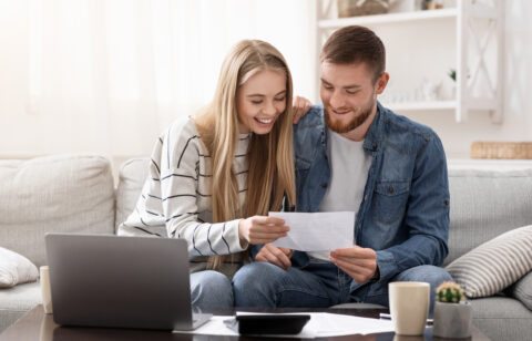 Couple smiling while reviewing paperwork together on a couch with a laptop.