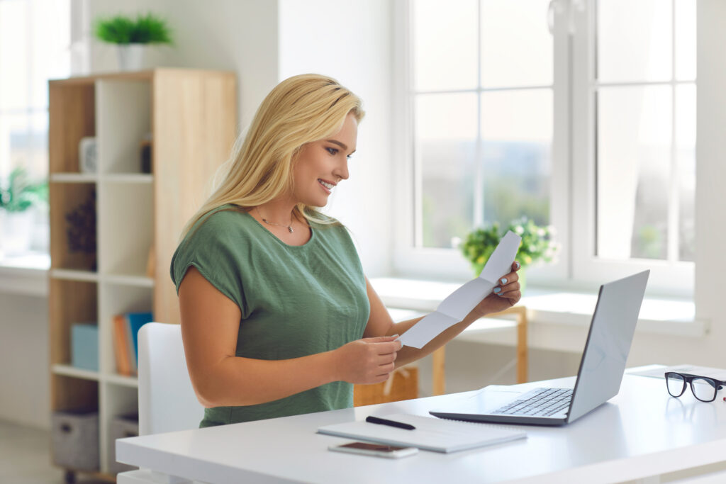 Woman smiling while reviewing paperwork at a desk with a laptop.