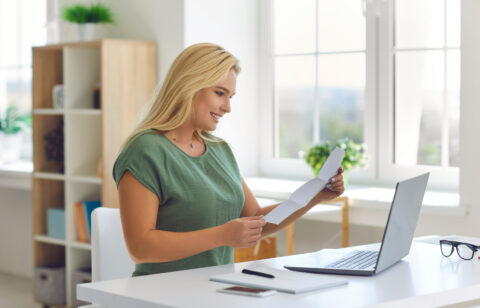 Woman smiling while reviewing paperwork at a desk with a laptop.