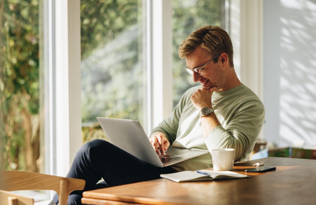 Smiling man using a laptop while sitting at a table in a sunlit room.