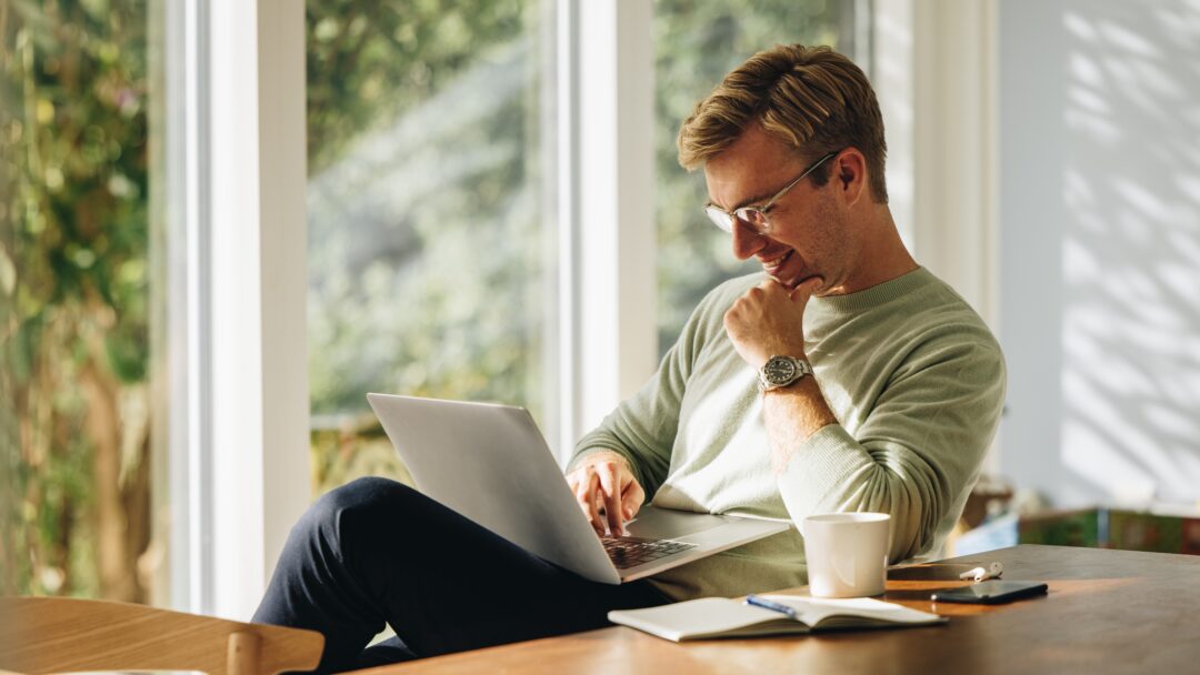 Smiling man using a laptop while sitting at a table in a sunlit room.