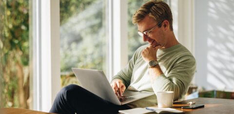 Smiling man using a laptop while sitting at a table in a sunlit room.