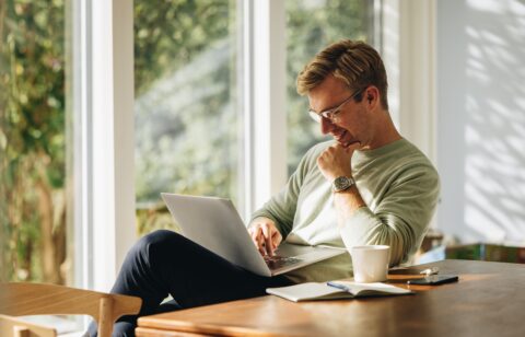 Smiling man using a laptop while sitting at a table in a sunlit room.