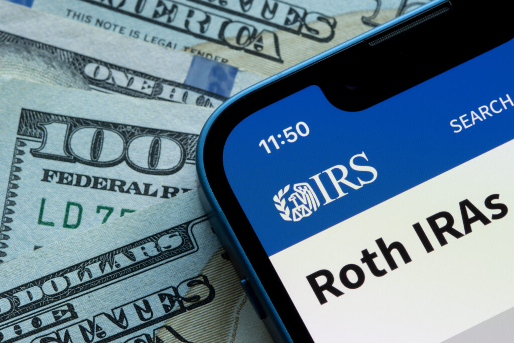 Smartphone displaying an IRS Roth IRA webpage beside U.S. dollar bills.
