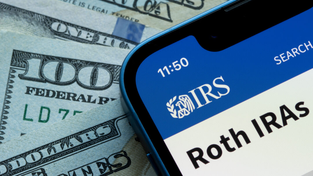 Smartphone displaying an IRS Roth IRA webpage beside U.S. dollar bills.