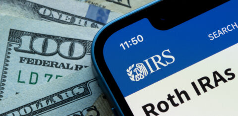 Smartphone displaying an IRS Roth IRA webpage beside U.S. dollar bills.