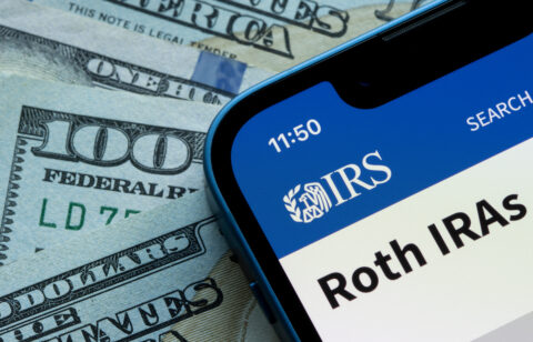 Smartphone displaying an IRS Roth IRA webpage beside U.S. dollar bills.