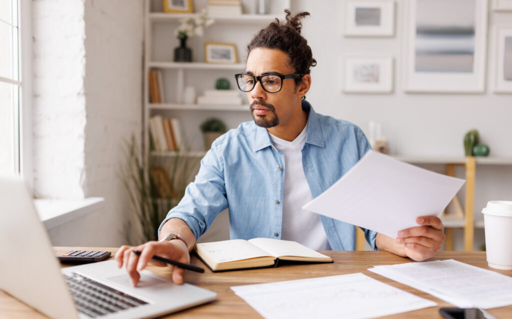 Man reviewing paperwork while working on a laptop at a desk.