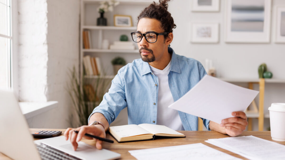Man reviewing paperwork while working on a laptop at a desk.