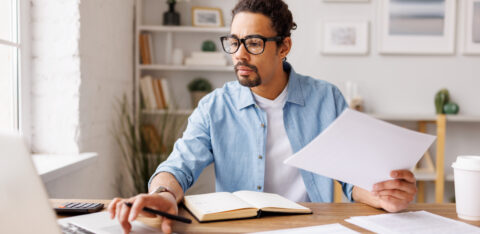 Man reviewing paperwork while working on a laptop at a desk.