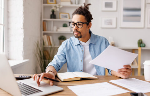 Man reviewing paperwork while working on a laptop at a desk.
