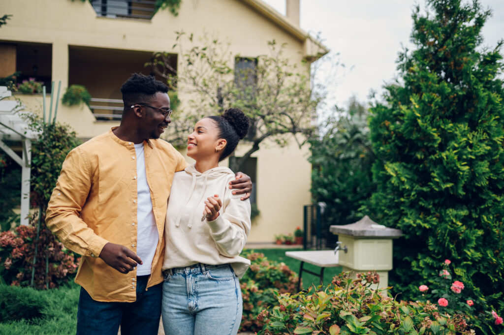 A smiling couple stands arm in arm outside a house surrounded by trees and flowers.