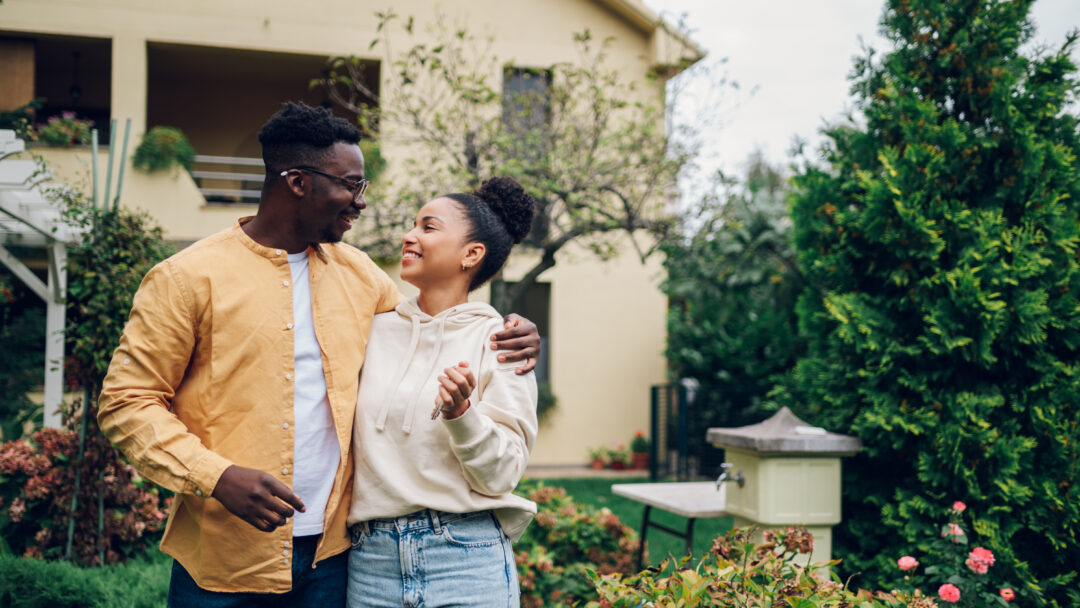 A smiling couple stands arm in arm outside a house surrounded by trees and flowers.