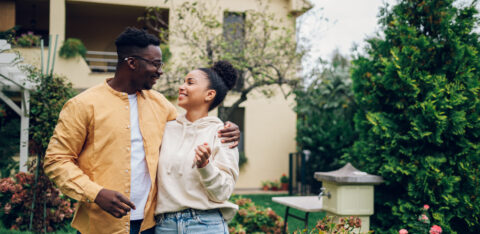 A smiling couple stands arm in arm outside a house surrounded by trees and flowers.