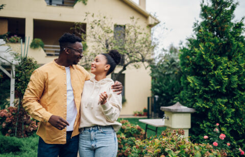 A smiling couple stands arm in arm outside a house surrounded by trees and flowers.