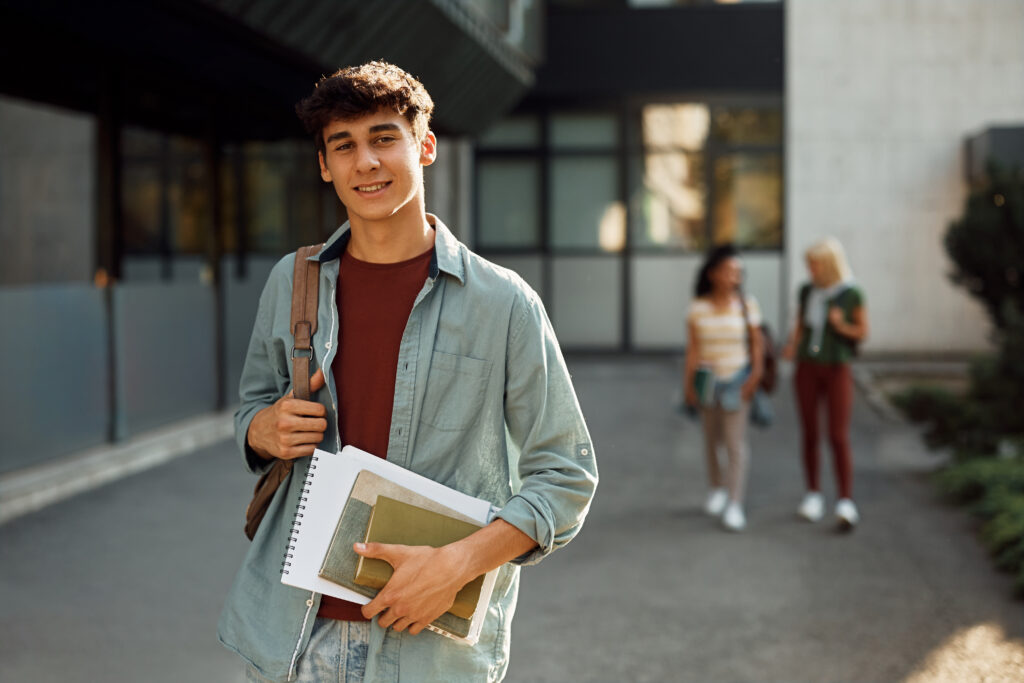 College student carrying books and a backpack while standing outside on campus.