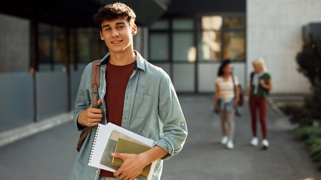 College student carrying books and a backpack while standing outside on campus.
