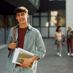 College student carrying books and a backpack while standing outside on campus.