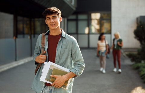 College student carrying books and a backpack while standing outside on campus.