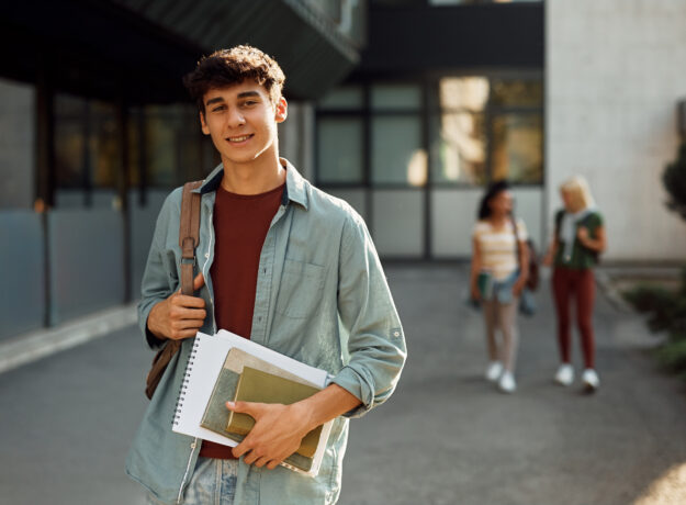 College student carrying books and a backpack while standing outside on campus.