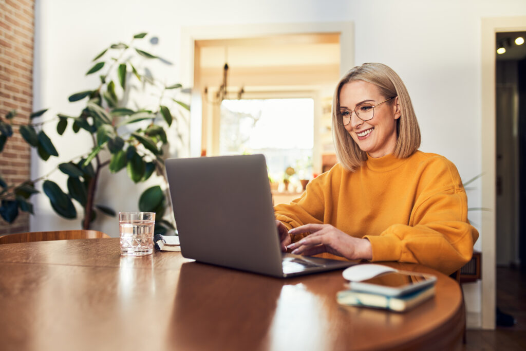 Smiling woman wearing glasses using a laptop while sitting at a wooden table at home.