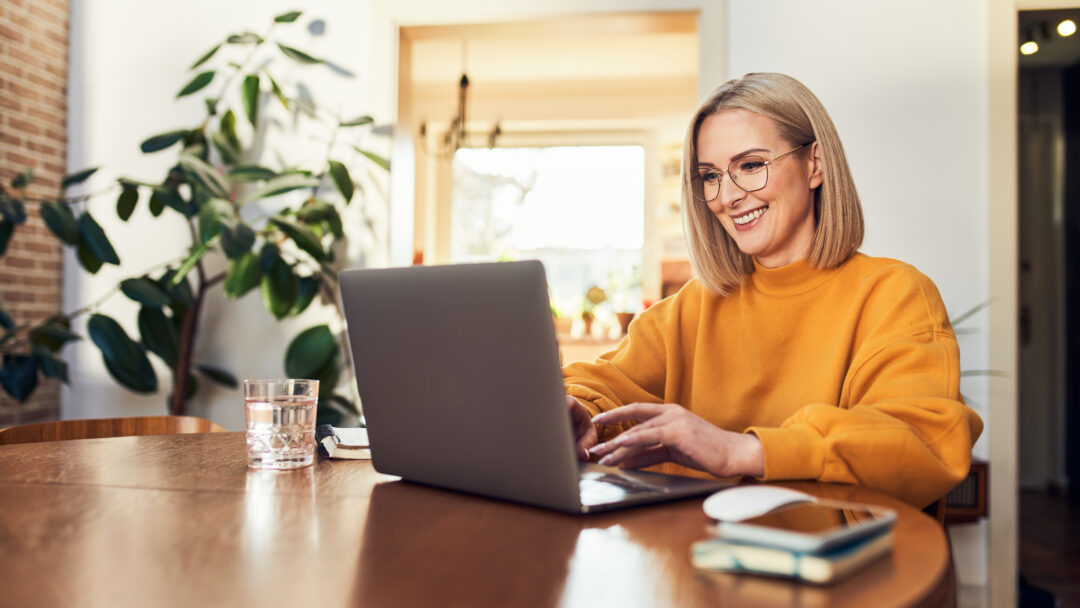 Smiling woman wearing glasses using a laptop while sitting at a wooden table at home.