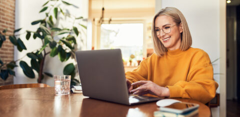Smiling woman wearing glasses using a laptop while sitting at a wooden table at home.