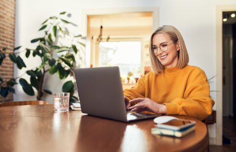 Smiling woman wearing glasses using a laptop while sitting at a wooden table at home.
