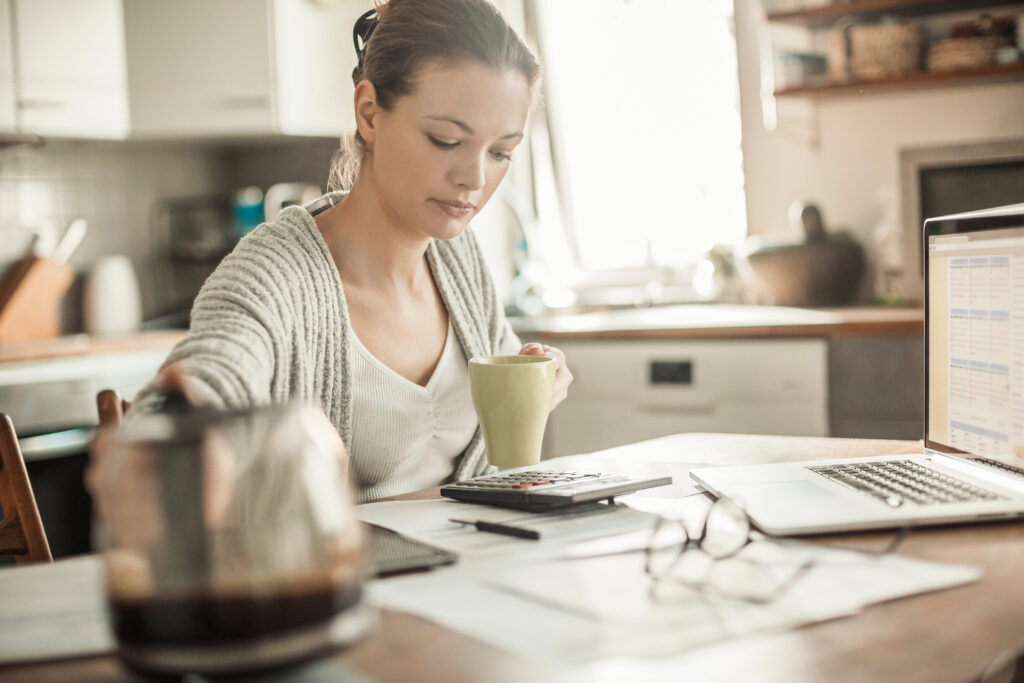 Young woman reviewing paperwork with a calculator and laptop at a kitchen table.