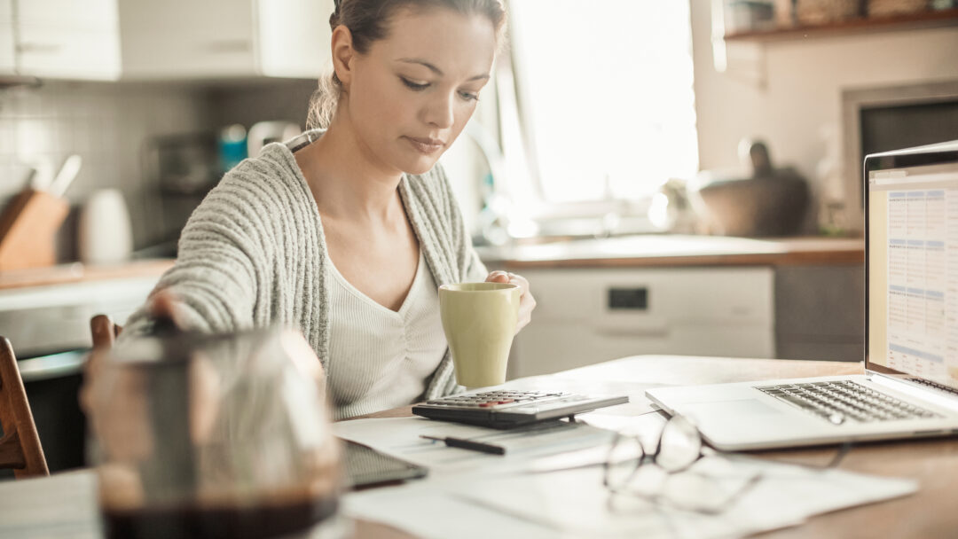 Young woman reviewing paperwork with a calculator and laptop at a kitchen table.