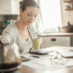 Young woman reviewing paperwork with a calculator and laptop at a kitchen table.
