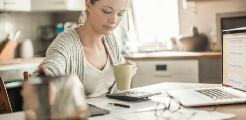 Young woman reviewing paperwork with a calculator and laptop at a kitchen table.