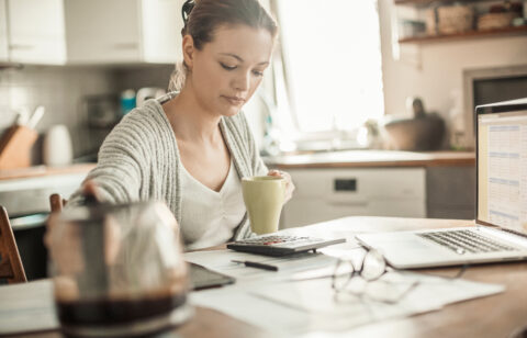 Young woman reviewing paperwork with a calculator and laptop at a kitchen table.
