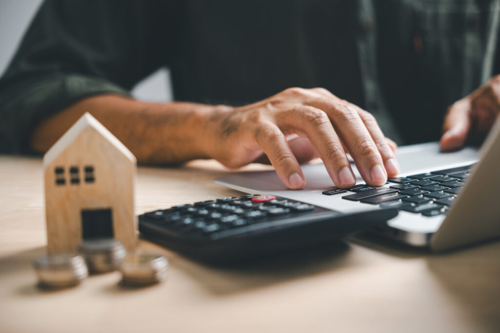 Person using a laptop beside a calculator, coins and a small wooden house.