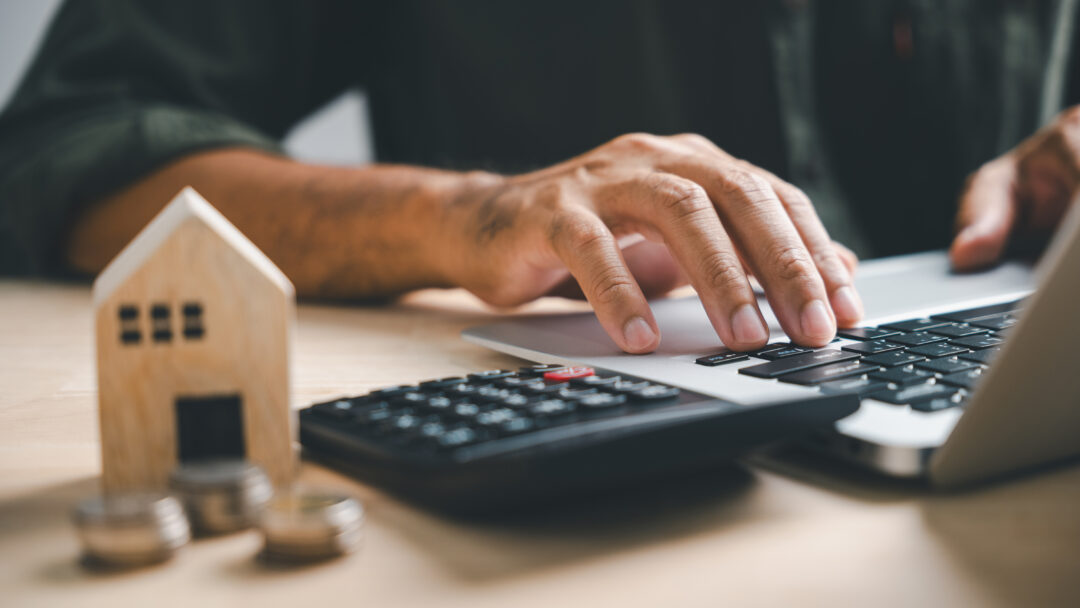 Person using a laptop beside a calculator, coins and a small wooden house.