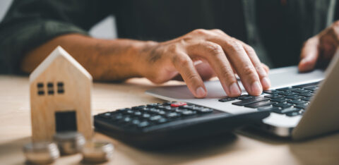 Person using a laptop beside a calculator, coins and a small wooden house.