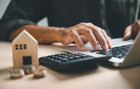 Person using a laptop beside a calculator, coins and a small wooden house.