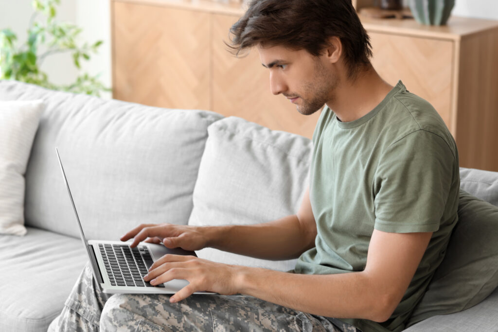 Young man in a green T-shirt and camouflage pants typing on a laptop on a sofa.