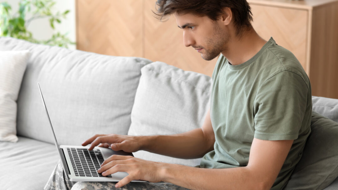Young man in a green T-shirt and camouflage pants typing on a laptop on a sofa.