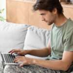 Young man in a green T-shirt and camouflage pants typing on a laptop on a sofa.