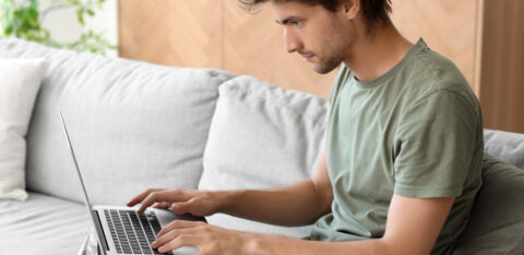 Young man in a green T-shirt and camouflage pants typing on a laptop on a sofa.