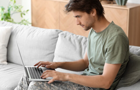 Young man in a green T-shirt and camouflage pants typing on a laptop on a sofa.