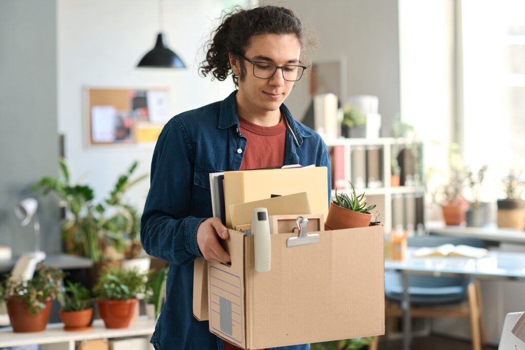 Young man in glasses carrying a box of personal items through an office.