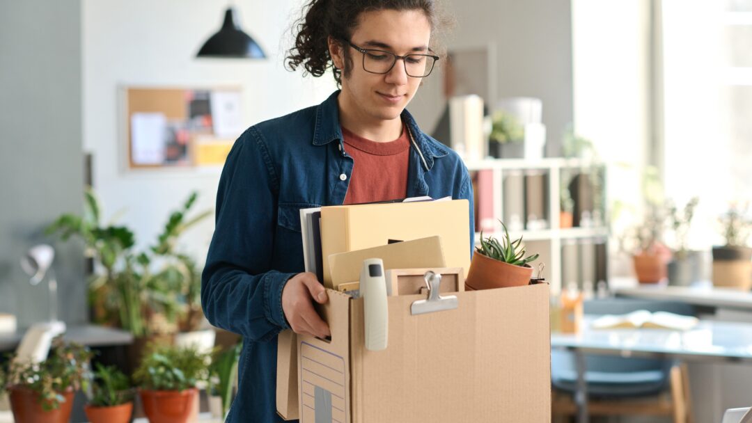 Young man in glasses carrying a box of personal items through an office.
